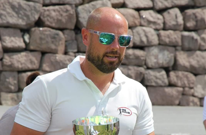 En mann med skjegg iført solbriller og en hvit poloskjorte som sitter utendørs, muligens på et sports- eller sosialt arrangement, med et snev av tilfredshet mens han lener seg mot et trofé på et bord. I