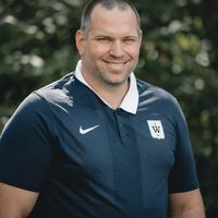 En mann, identifisert som Fredrik Aukland, smilende til kamera, ikledd mørk poloskjorte med logo på venstre side, med uskarpt grønt i bakgrunnen.
