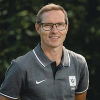 En smilende mann med briller og krageskjorte med Asgeir Fremstad-logo, stående utendørs med grønt i bakgrunnen.
