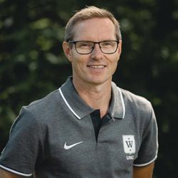 En smilende mann med briller og krageskjorte med Asgeir Fremstad-logo, stående utendørs med grønt i bakgrunnen.