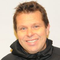 En mann med et varmt smil iført svart hettegenser mot lys bakgrunn er Mikael Flygind Larsen.