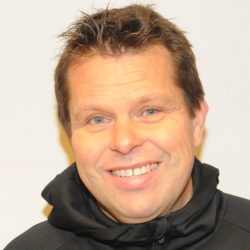 En mann med et varmt smil iført svart hettegenser mot lys bakgrunn er Mikael Flygind Larsen.