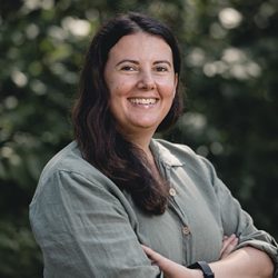 En smilende kvinne med mørkt hår, iført en uformell grønn skjorte, står selvsikkert utendørs med armene i kors, og minner om stilen Fredrik Aukland fanget.