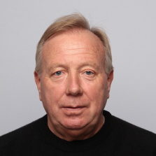 Et hodebilde av Mikael Flygind Larsen, en middelaldrende mann med lys hud, med kort blondt hår, og iført en svart topp, mot en lysegrå bakgrunn.