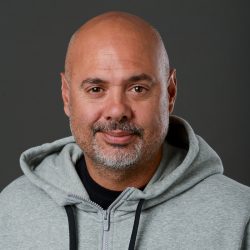 En selvsikker, Christopher Twiddy, med skjeggstubbe, iført en grå hettegenser, smiler forsiktig til kameraet mot en mørk bakgrunn.