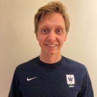 En smilende person med kort blondt hår iført mørk Espen Honganvik Holen sportsskjorte med logo og årstallet 1907 på.
