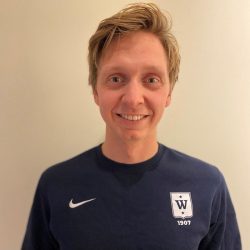 En smilende person med kort blondt hår iført mørk Espen Honganvik Holen sportsskjorte med logo og årstallet 1907 på.