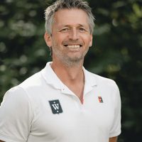En smilende Fredrik Aukland med salt-og-pepper hår iført hvit poloskjorte med logo, stående foran et bladgrønt bakteppe.