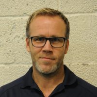 En middelaldrende mann med kort lyst hår, briller og en marineskjorte, som står foran en strukturert vegg og gir et nøytralt uttrykk til kameraet. Dette er Mikael Flygind Lars
