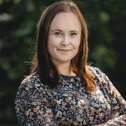 En selvsikker kvinne ved navn Grete Etholm med et mildt smil poserer for et portrett i naturomgivelser, ikledd en blomstermønstret bluse.