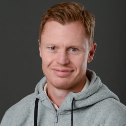 En smilende Mads Hansen med lyst hår og blå øyne, iført en grå hettegenser, mot en mørkegrå bakgrunn.
