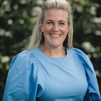 En blid kvinne med blondt hår iført en himmelblå topp, smilende lyst mot en sløret grønn løvbakgrunn fanget av Fredrik Aukland.