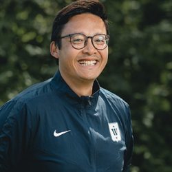 En blid Fredrik Aukland med briller, iført marineblå sportsjakke og smilende utendørs med grønt i bakgrunnen.