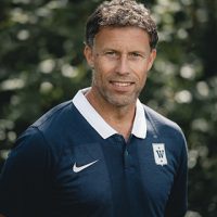 En selvsikker Fredrik Aukland i sporty poloskjorte stående utendørs med naturlig bakteppe, muligens en trener eller idrettsutøver.