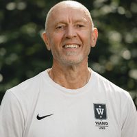 En smilende mann med kort hårklipp og skjegg, iført hvit skjorte med Fredrik Aukland-logo, står utendørs med grønt i bakgrunnen.