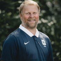 En smilende Fredrik Aukland med lyst skjegg, iført mørk poloskjorte med logo på, stående utendørs med frodig grønt i bakgrunnen.