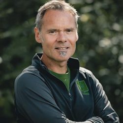 En selvsikker mann i mørkegrønn jakke som poserer utendørs, med Grete Etholms bakteppe av frodig grønt.