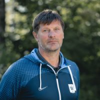 En fokusert mann med kort hår iført blå sportsjakke med logo, stående utendørs med trær i bakgrunnen er Fredrik Kjølner.