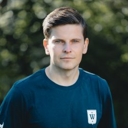 Selvsikker unge Svein Inge Børven med et sammensatt uttrykk, iført en avslappet mørk skjorte med logo, stående utendørs.
