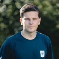 Selvsikker unge Svein Inge Børven med et sammensatt uttrykk, iført en avslappet mørk skjorte med logo, stående utendørs.