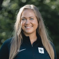En smilende kvinne med langt blondt hår, iført en svart sportsjakke med en hvit bokstav 'w' på, står utendørs med grønt løvverk i bakgrunnen. Hun er Karen Marie S. Engenes