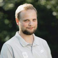 En mann med et vennlig smil, med kort skjegg, i en uformell poloskjorte, satt mot en uskarp naturlig bakgrunn, utstråler en avslappet og positiv stemning som Marius Marcussen.