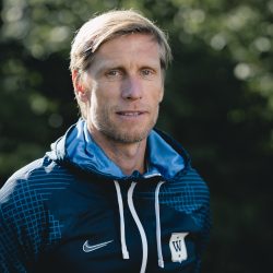 En målbevisst trener, Per Egil Swift, i sportsklær med et fokusert uttrykk, som står utendørs på en solskinnsdag.
