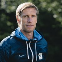 En målbevisst trener, Per Egil Swift, i sportsklær med et fokusert uttrykk, som står utendørs på en solskinnsdag.