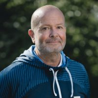En selvsikker, middelaldrende mann med skjegg, iført en blå hettegenser, smiler forsiktig mens han står utendørs. Dette er ingen ringere enn Tor Øyvind Berntsen.