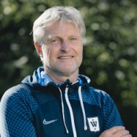 En selvsikker mann med en vennlig oppførsel, iført en uformell sportsjakke og et subtilt smil, står utendørs med løvverk i bakgrunnen og gir fra seg en atmosfære av tilgjengelighet og profesjonalitet.