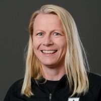 En smilende kvinne med langt blondt hår, lyse øyne og en svart topp mot grå bakgrunn er Heidi Fageraas Lock.