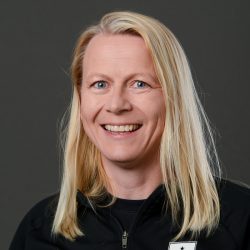 En smilende kvinne med langt blondt hår, lyse øyne og en svart topp mot grå bakgrunn er Heidi Fageraas Lock.