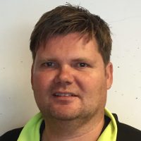 Et nærbildeportrett av Andre Jauert med kort hår, med et nøytralt uttrykk mot en vanlig bakgrunn, iført det som ser ut til å være en svart og fluorescerende limejakke eller vest.