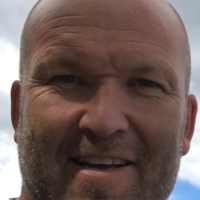 Nærbildeportrett av Andreas Holter smilende med barbert hode mot et bakteppe av en overskyet himmel.
