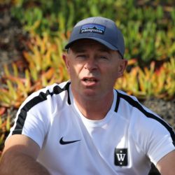 En mann med baseballcaps med Anker Frithjof Thomsen-logoen og en svart-hvit sportsskjorte ser ut til å være midt i samtalen eller gi instruksjoner utendørs, med bakgrunn
