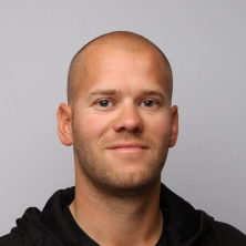 En mann med barbert hode og et snev av et smil, iført en svart topp, poserer for et hodeskudd mot en lysegrå bakgrunn.
