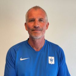 En smilende mann i blå sportsskjorte med hvit logo på brystet, identifisert som Atle Mortvedt, stående mot en vanlig bakgrunn.