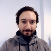 En smilende person, Christian Lopez Salas, med skjegg og bart, iført en grå hettegenser, mot en vanlig bakgrunn.