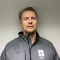 En mann med lys skjeggstubb stående foran en vanlig bakgrunn, iført en grå glidelåsjakke med Even Ramsdal-logoen på venstre side.