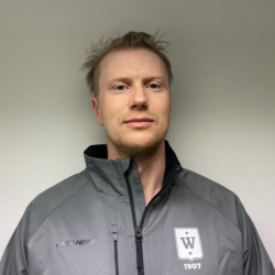 En mann med lys skjeggstubb stående foran en vanlig bakgrunn, iført en grå glidelåsjakke med Even Ramsdal-logoen på venstre side.