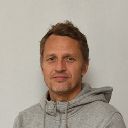 En uformelt kledd mann med et lett smil, identifisert som Flemming Solberg, iført en grå hettegenser, mot ensfarget bakgrunn.