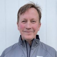 En smilende mann med kort hår, iført en grå jakke, stående mot en vanlig hvit bakgrunn.