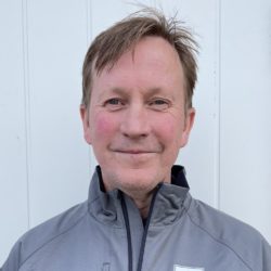 En smilende mann med kort hår, iført en grå jakke, stående mot en vanlig hvit bakgrunn.