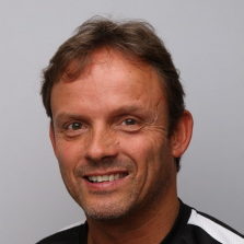 En mann med et vennlig smil, kort hår og skjeggstubber på grå bakgrunn er Håkon Henne.