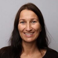 Et portrett av Heidi Uldal smilende, med langt mørkt hår og svart topp mot en nøytral grå bakgrunn.