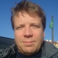 En mann med kort hår og et subtilt smil, Frank Nordseth nyter utendørs på en solrik dag med klarblå himmel i bakgrunnen.