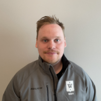 En selvsikker mann med kammet hår, med bart og lyst skjegg, iført en grå genser med kvart glidelås med Kim Veisten-logoen på, poserer for et bilde mot en