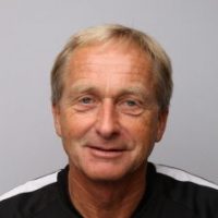 Et portrett av en smilende middelaldrende mann ved navn Kjetil Østbø med kort blondt hår, iført svart topp mot grå bakgrunn.