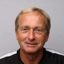 Et portrett av en smilende middelaldrende mann ved navn Kjetil Østbø med kort blondt hår, iført svart topp mot grå bakgrunn.