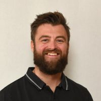 En munter skjeggete mann ved navn Kristian Gilbert, med et varmt smil, iført svart skjorte med krage mot ensfarget bakgrunn.